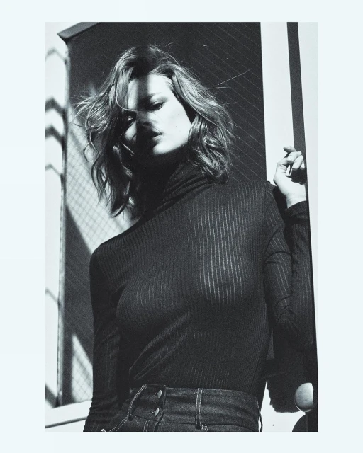 Black and white portrait of a woman with shoulder-length wavy hair wearing a black turtleneck and jeans, posing near a window.
