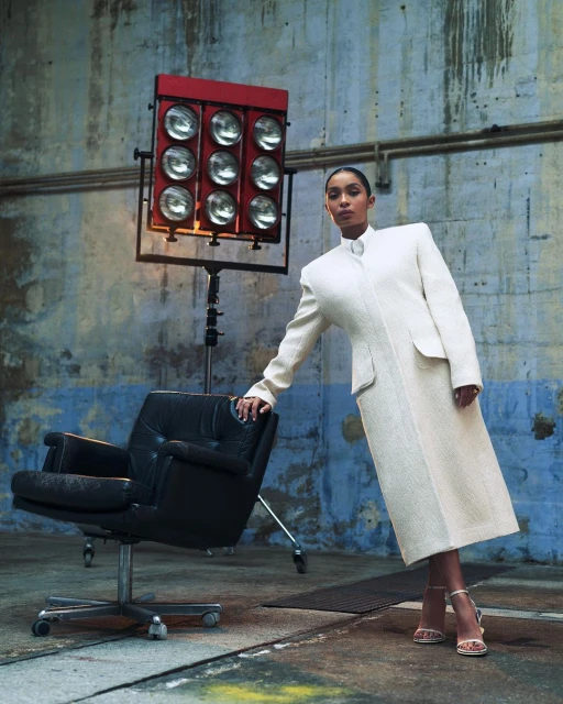 Elegant woman in a cream coat poses with an armchair and a spotlight in an industrial photo studio with textured concrete walls.