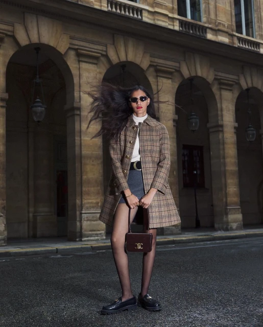 Woman in plaid coat, mini skirt, and tights with a purse stands on a city street in front of building with arched openings.