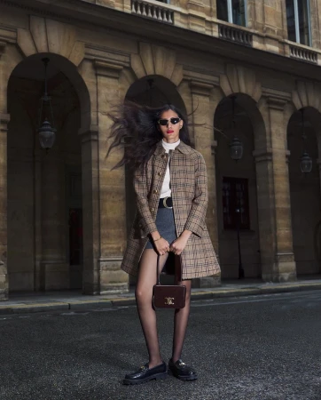 Woman in plaid coat, mini skirt, and tights with a purse stands on a city street in front of building with arched openings.