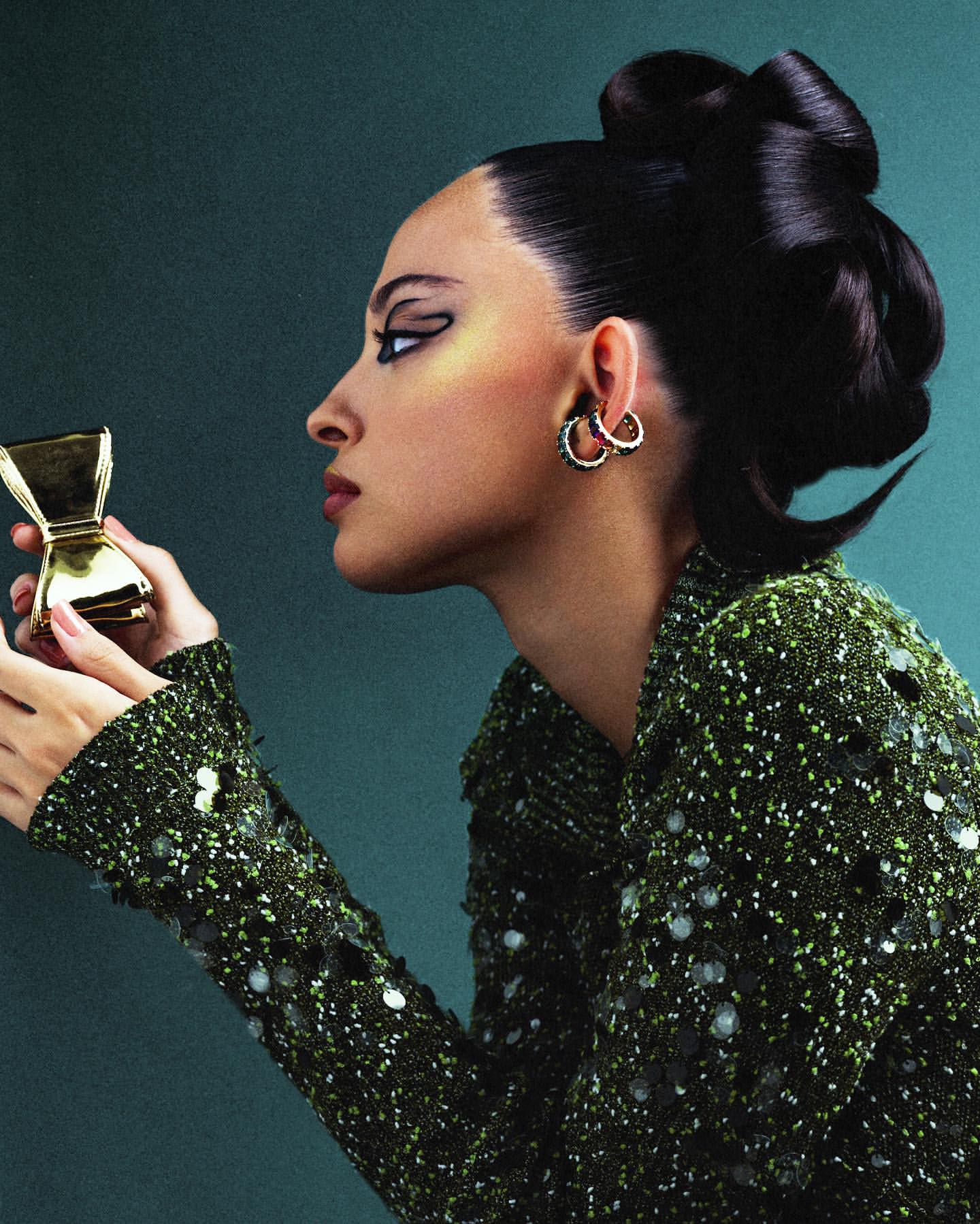 Woman in green sequined jacket holds a gold hourglass against a teal backdrop, showcasing dramatic eye makeup and an elegant updo hairstyle.