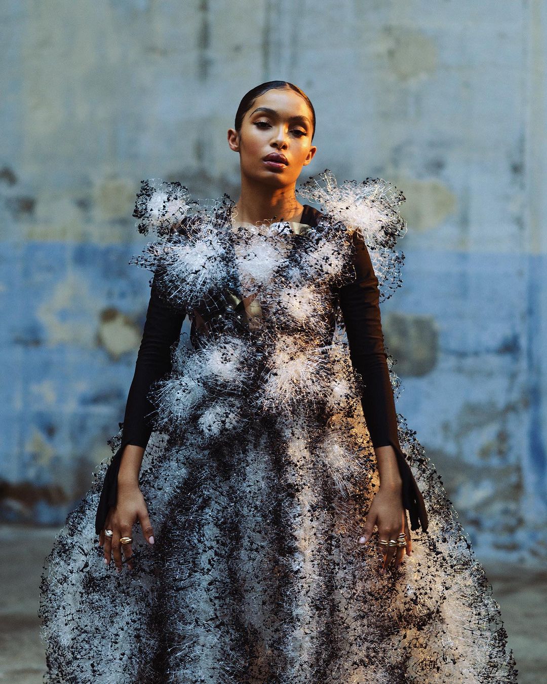 Woman in an avant-garde black and white dress made of thread stands against a textured wall in a fashion forward portrait.