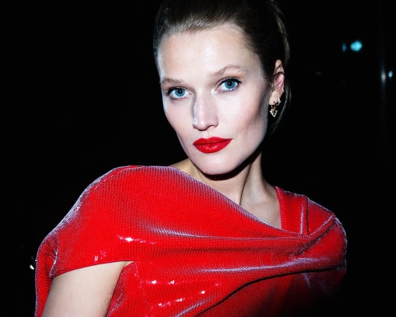 Close-up portrait of a woman with fair skin, red lips, and blue eyes wearing a draped red sequined top against a dark background.