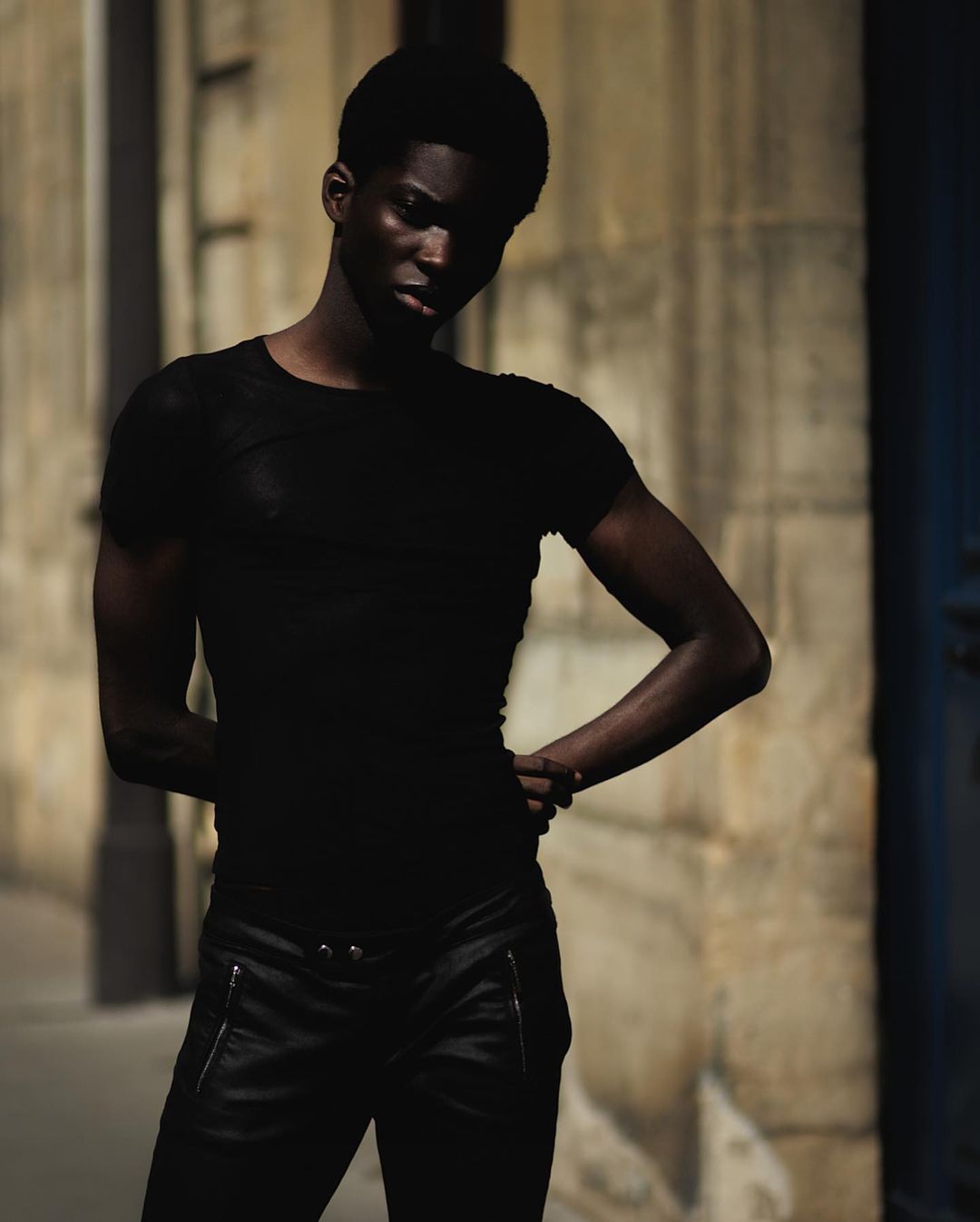 Black man wearing black t-shirt and dark trousers posing outdoors against stone wall by Rayan Ayash
