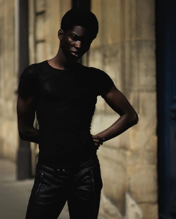 Black man wearing black t-shirt and dark trousers posing outdoors against stone wall by Rayan Ayash