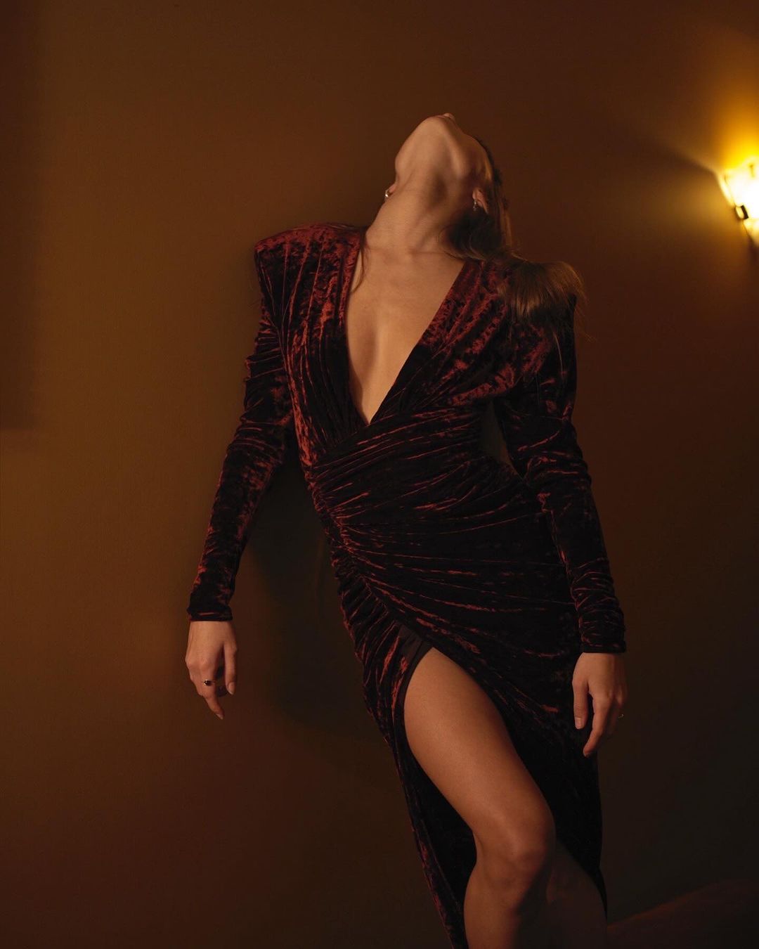 Woman in a long-sleeve velvet dress with a thigh-high slit, leaning back against a wall under warm lighting. Fashion photography.