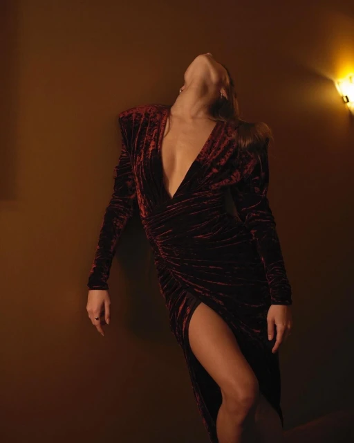 Woman in a long-sleeve velvet dress with a thigh-high slit, leaning back against a wall under warm lighting. Fashion photography.