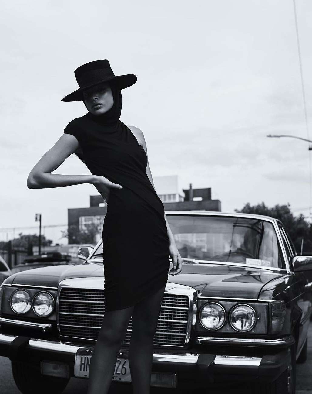 Monochrome fashion portrait of a stylish adult female wearing a black dress and hat next to a classic car on an urban street.