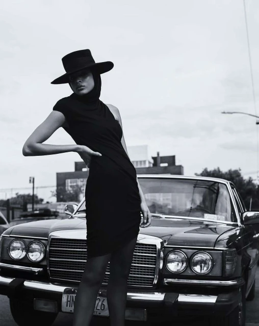 Monochrome fashion portrait of a stylish adult female wearing a black dress and hat next to a classic car on an urban street.