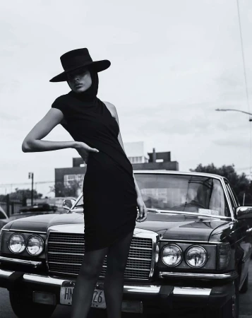 Monochrome fashion portrait of a stylish adult female wearing a black dress and hat next to a classic car on an urban street.