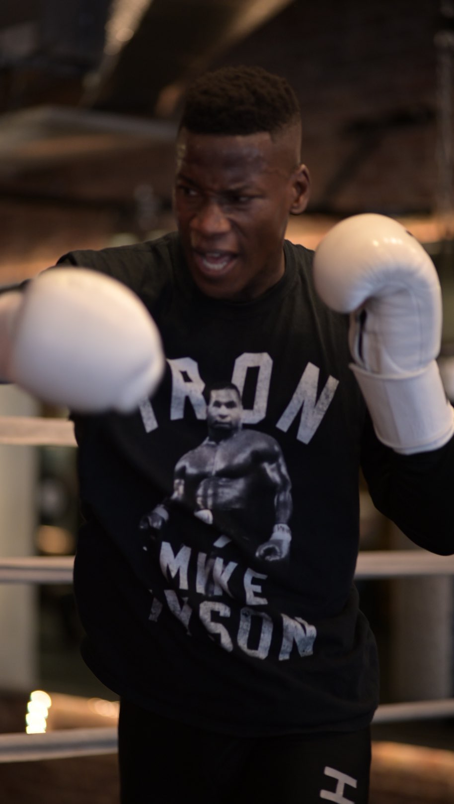 Boxer in a black t-shirt with Mike Tyson graphic, wearing white boxing gloves, in a boxing ring with a neutral interior background.