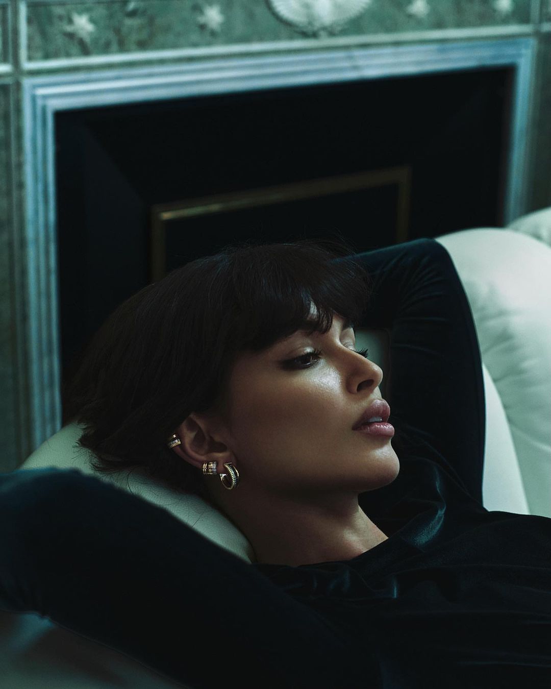 Close-up profile of a woman with dark hair and bangs reclining on a white couch, wearing gold hoop earrings and ear cuffs.
