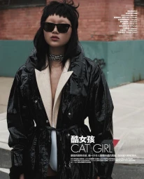 Model with dark sunglasses wearing a glossy black vinyl coat outdoors by a red brick building