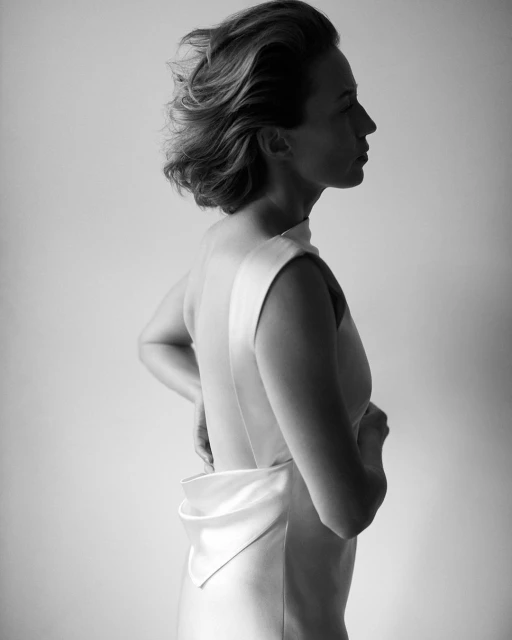 Black and white portrait of a woman in a light-colored dress with open back, posing in a studio setting, with soft lighting.
