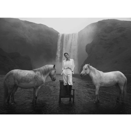 A person standing on a chair in white attire flanked by two white horses with a large waterfall in the background