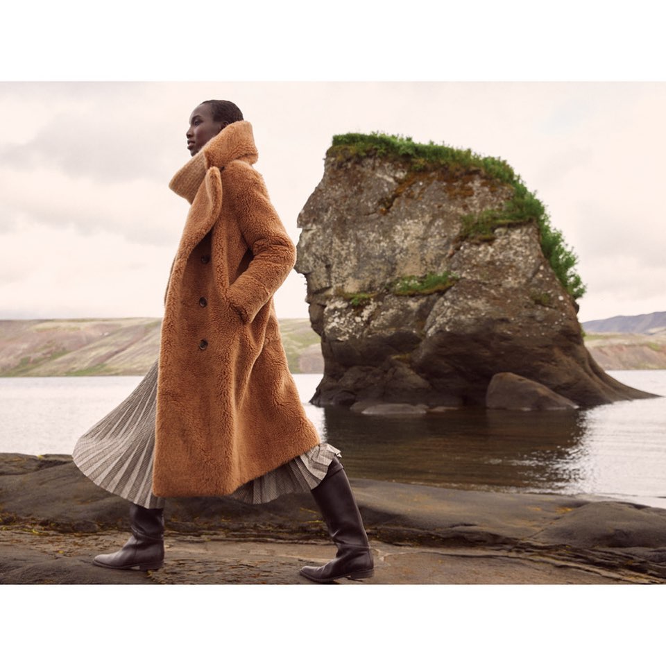 Model in a brown shearling coat and pleated skirt walking past a large rock formation near water.