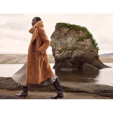 Model in a brown shearling coat and pleated skirt walking past a large rock formation near water.