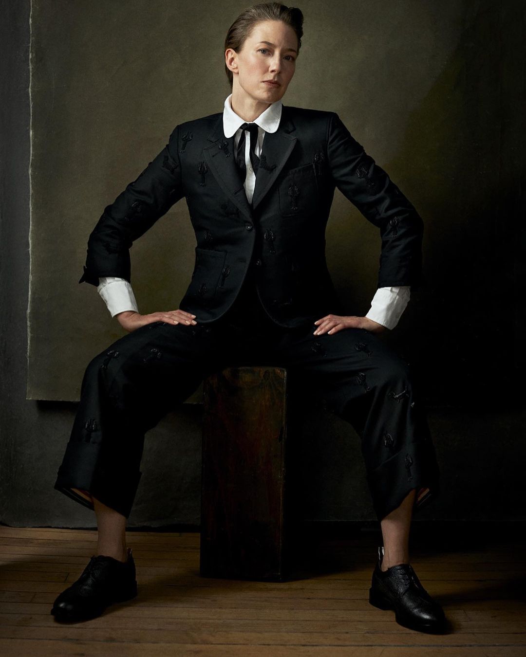 Portrait of a woman in a black suit sitting on a wooden cube, looking at the camera in a studio setting with a neutral backdrop.