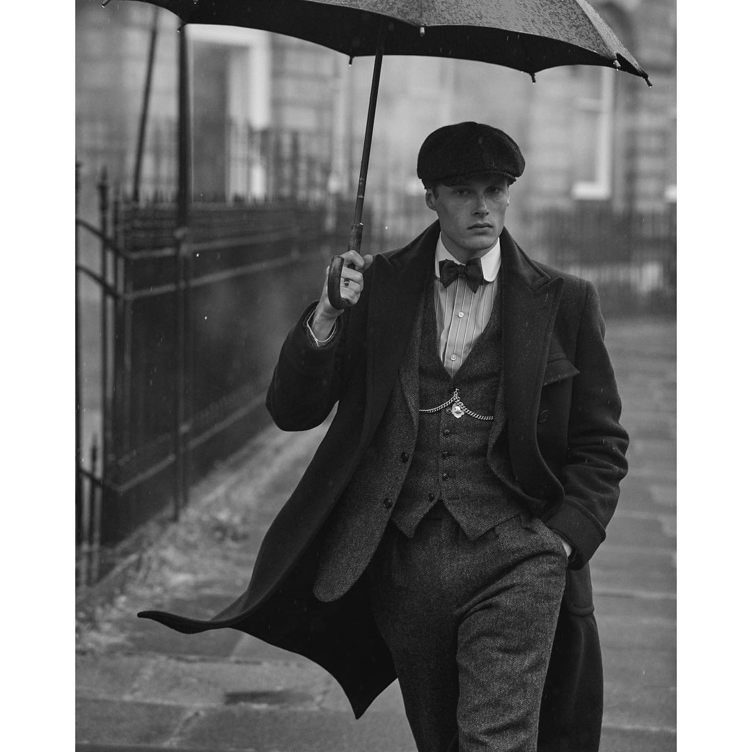 Black-and-white portrait of a man in a tweed suit and cap holding an umbrella in the rain on a city street.