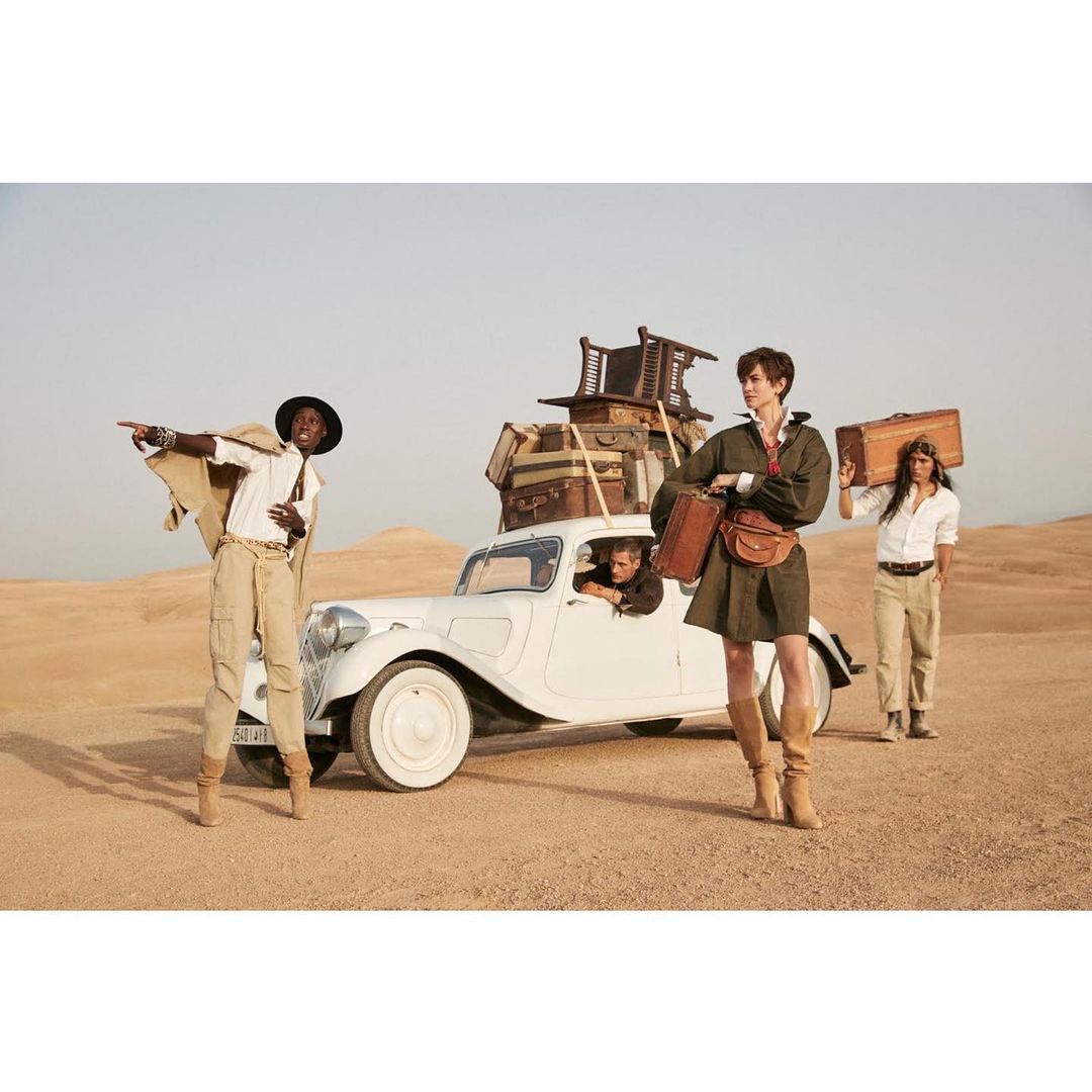 Four people and a driver stand near a vintage car in the desert with luggage piled on top, wearing stylish vintage-inspired outfits.