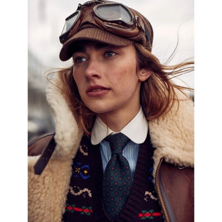 A young woman in an aviator hat and vintage-style clothing gazes off-camera.  She is wearing a shearling coat, shirt, and tie.