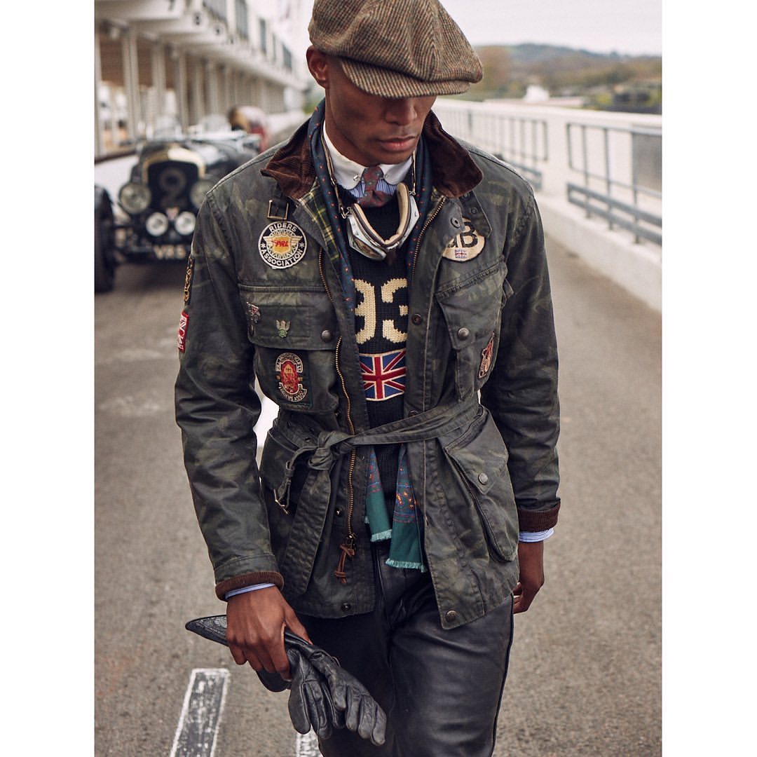 Stylish adult male model wears a vintage-inspired racing jacket, cap, and gloves, posing with a classic car in the background.