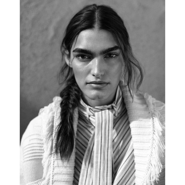 Monochrome portrait of a woman with braided hair, wearing a striped top and cardigan, against a neutral background. Fashion editorial.