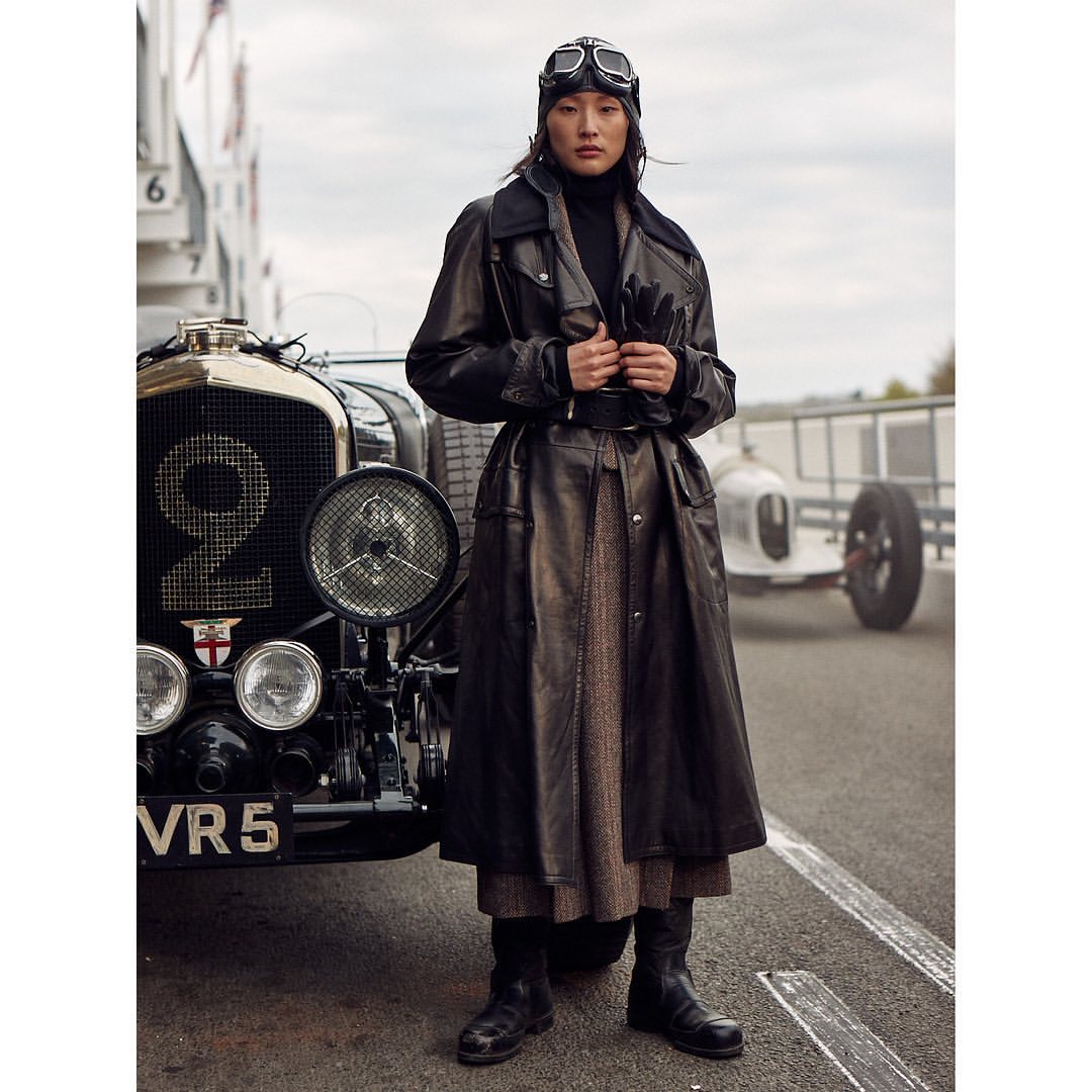 Woman in leather coat with vintage aviator goggles standing by a vintage Bentley race car on an outdoor track. Fashion editorial.