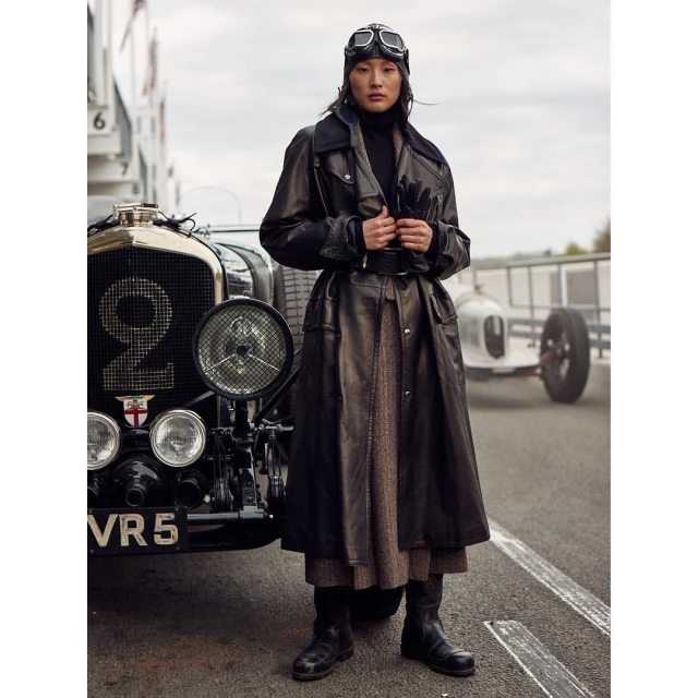 Woman in leather coat with vintage aviator goggles standing by a vintage Bentley race car on an outdoor track. Fashion editorial.