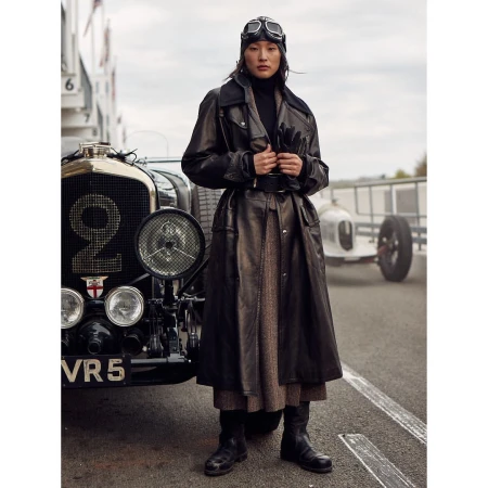 Woman in leather coat with vintage aviator goggles standing by a vintage Bentley race car on an outdoor track. Fashion editorial.