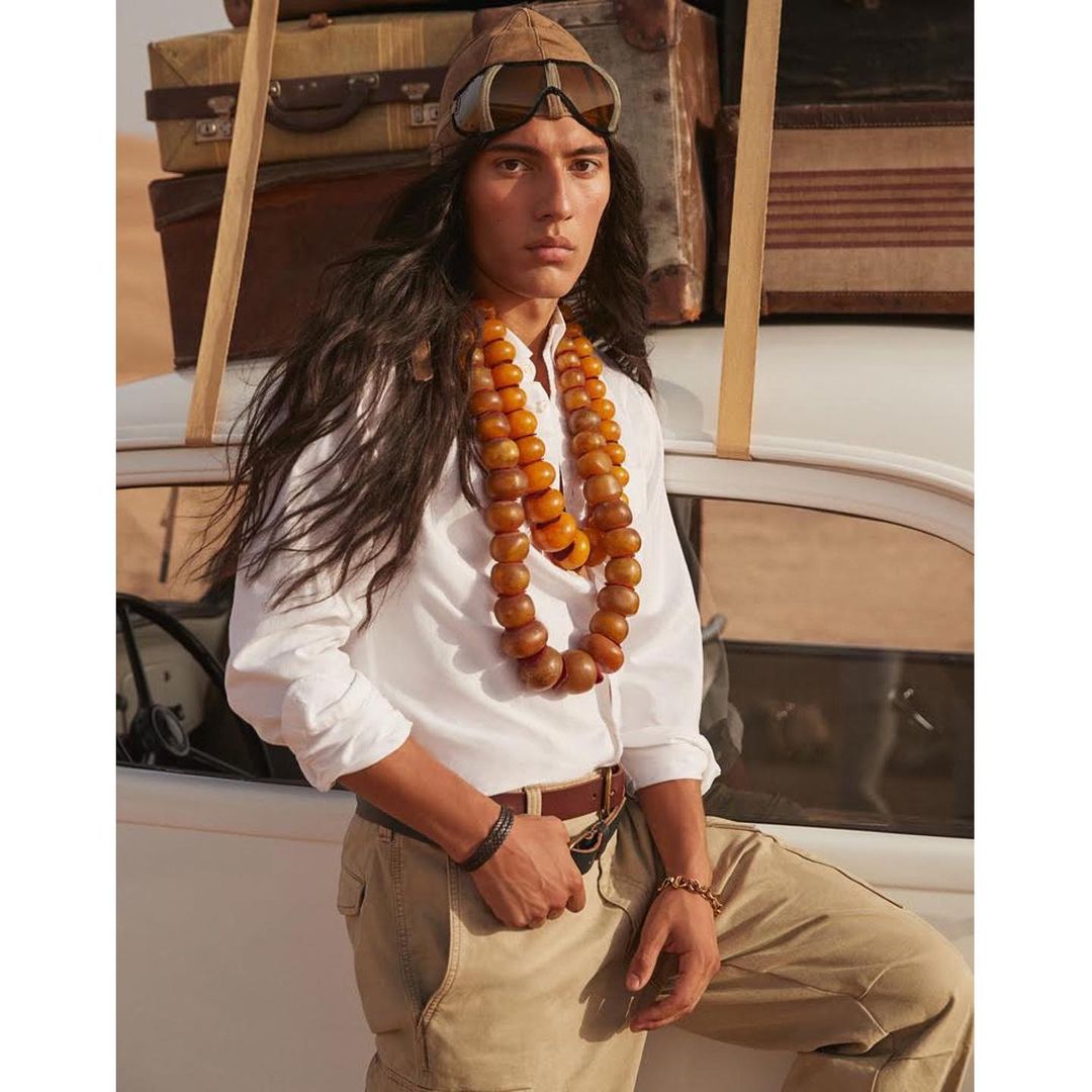 Man poses by car with stacked luggage, wearing a light shirt, khaki pants, cap, goggles, and beaded necklace in a desert setting.