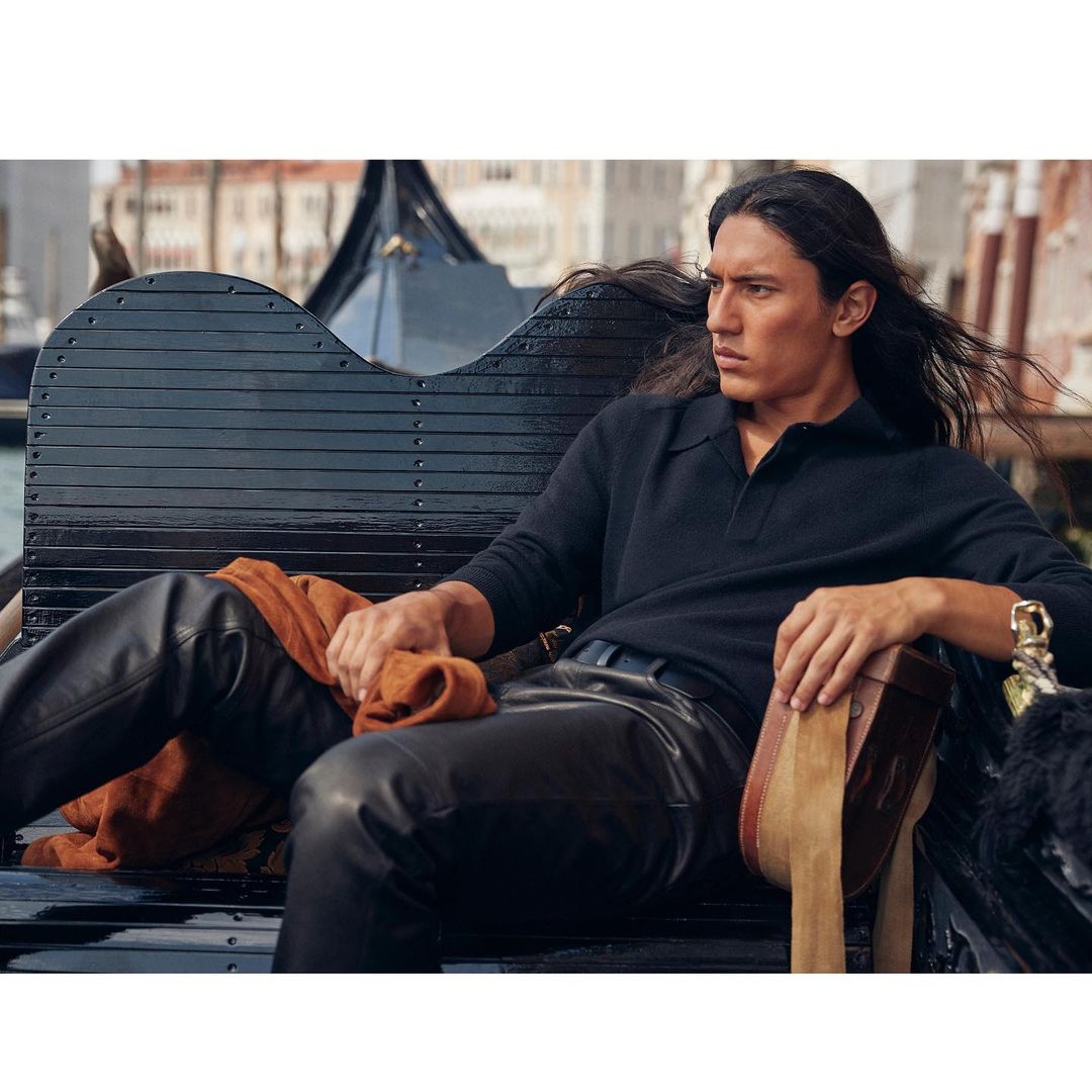 Dark-haired man in a black polo and black leather pants sits on a black gondola in Venice, Italy, enjoying the scenery.