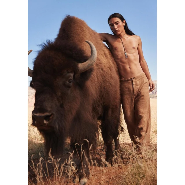 Eye-level shot of an Indigenous man with long hair standing next to a large bison in a grassy, arid landscape under a clear sky.