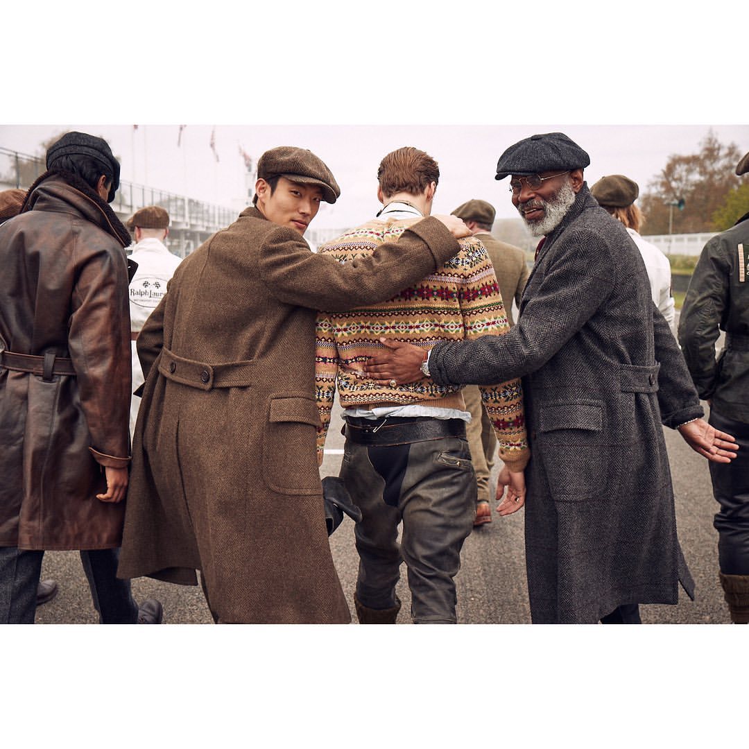 Three men in vintage coats and hats stand together on a racetrack, showcasing a blend of classic fashion and automotive culture.