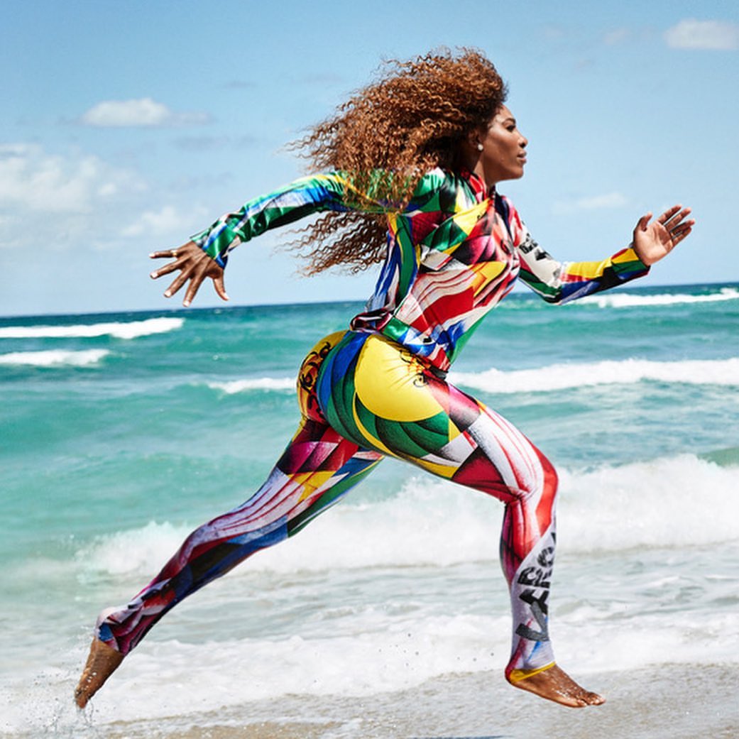 Athlete Serena Williams runs barefoot on a beach wearing a colorful, full-body suit with blue ocean water and a blue sky in the background.