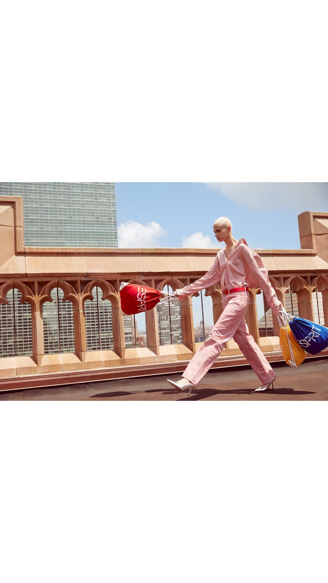 Fashion model walks on a rooftop in a pink outfit carrying drawstring bags with a cityscape and blue sky in the background.