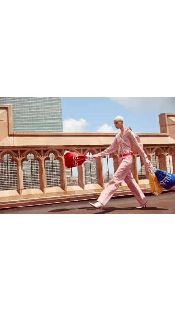 Fashion model walks on a rooftop in a pink outfit carrying drawstring bags with a cityscape and blue sky in the background.
