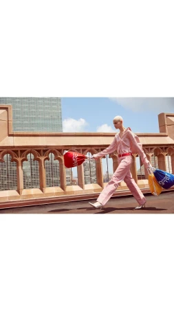 Fashion model walks on a rooftop in a pink outfit carrying drawstring bags with a cityscape and blue sky in the background.