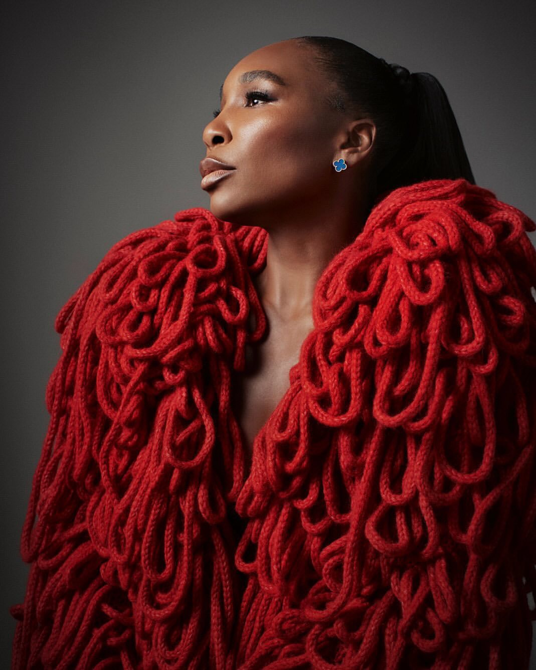 Portrait of an adult female with dark hair in a ponytail, posing in a bright red textured jacket against a dark grey backdrop.