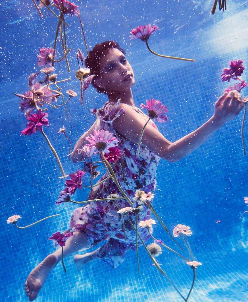 Woman in a floral dress underwater with gerbera daisies, blue mosaic tiles, and diffused natural light creating a dreamlike, ethereal scene.