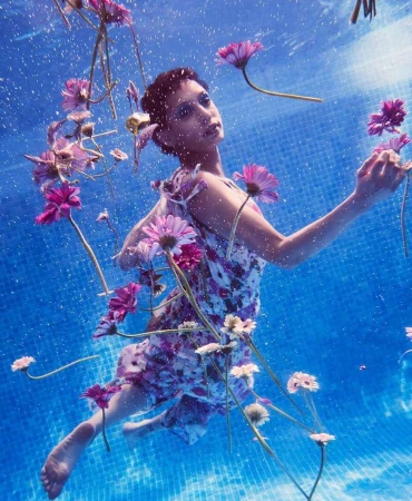 Woman in a floral dress underwater with gerbera daisies, blue mosaic tiles, and diffused natural light creating a dreamlike, ethereal scene.