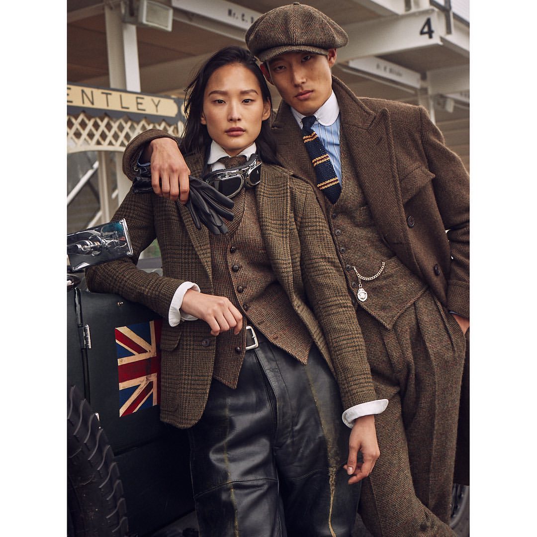 An Asian man and woman in vintage clothing, leaning against a car with a Union Jack, showcasing classic fashion in an outdoor setting.