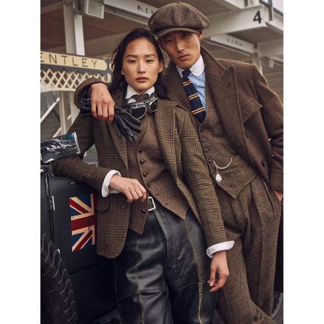 An Asian man and woman in vintage clothing, leaning against a car with a Union Jack, showcasing classic fashion in an outdoor setting.