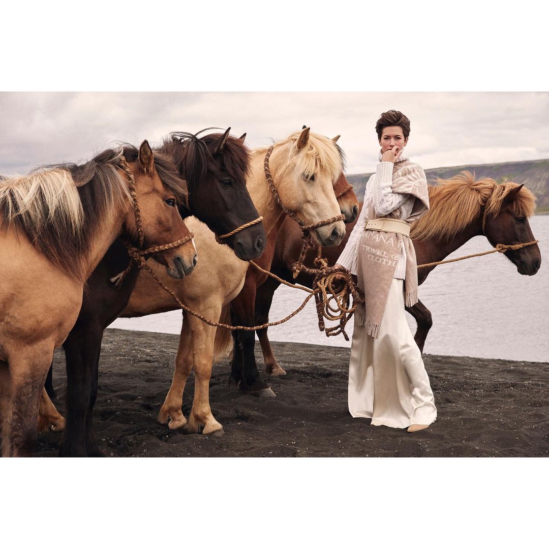 A woman in an elegant dress stands among five Icelandic horses on a black sand beach with a cloudy sky and water in the background.
