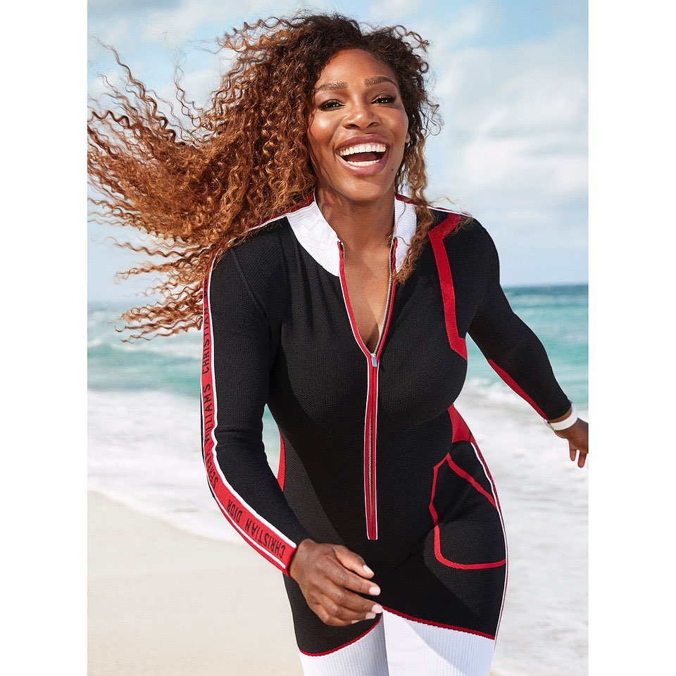 Serena Williams smiles on a beach wearing a black and white long-sleeved dress with a red zipper and lettering detail on the sleeves.