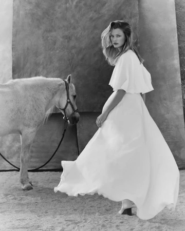 Monochrome fashion portrait of a woman in a white cape dress standing next to a white horse on sandy ground.