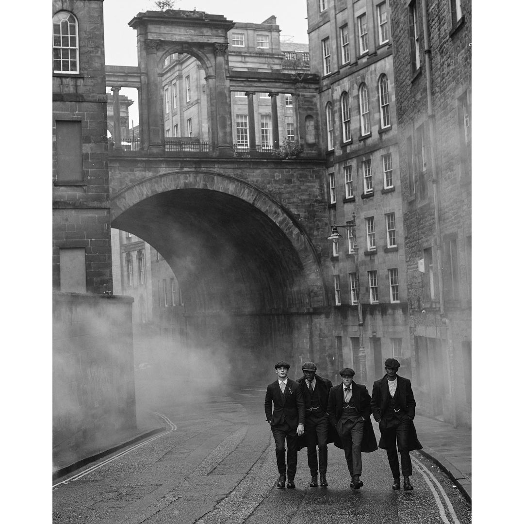 Four men in 1920s style attire walking on a cobblestone street under a stone bridge and buildings in a black and white photograph.