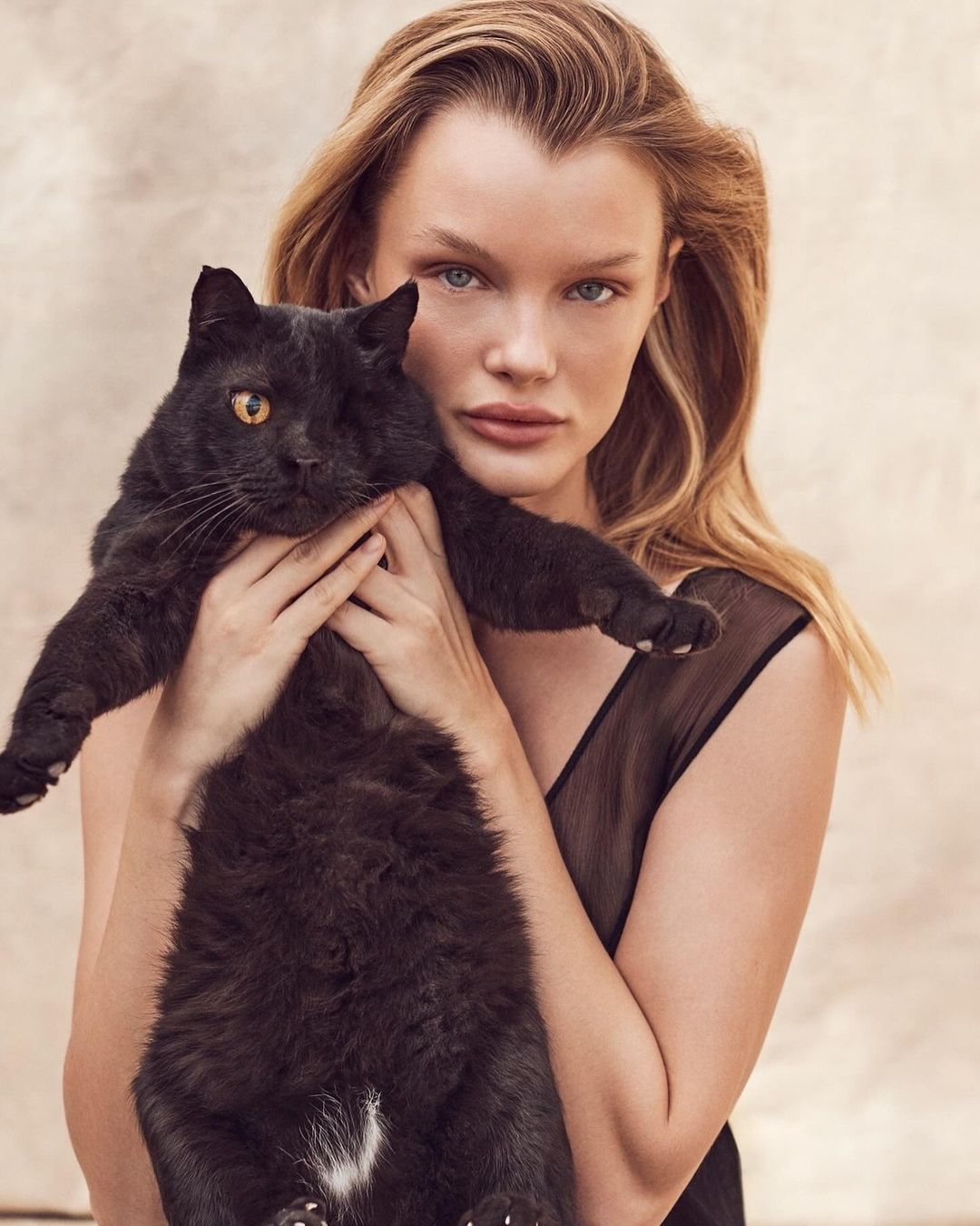 Portrait of a blonde adult woman holding a fluffy black cat with yellow eyes, in front of a neutral cream-colored background.
