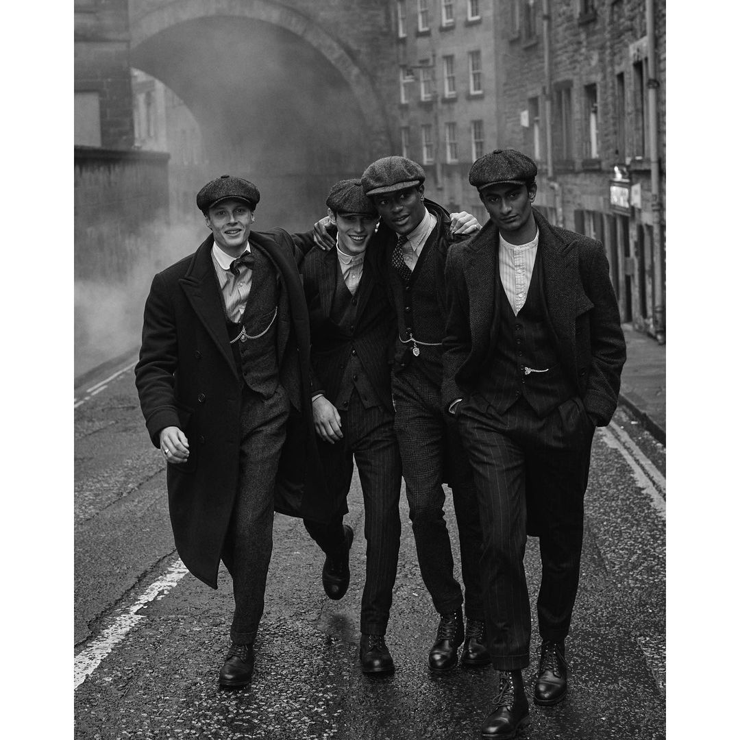 Four men in vintage attire walk on a cobblestone street beneath an archway, in a black and white outdoor fashion editorial shot.