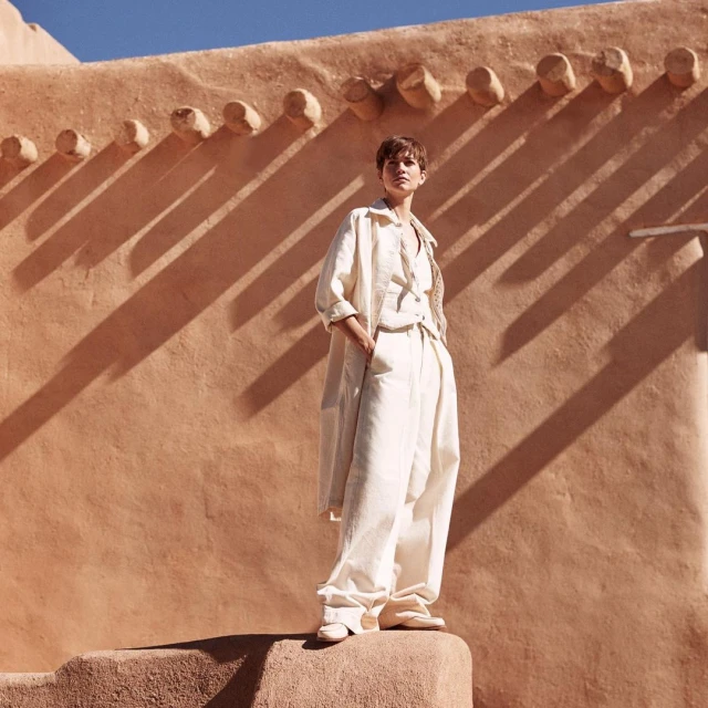 Woman in white linen outfit stands against a terracotta-colored building with geometric shadow patterns in soft, natural light.